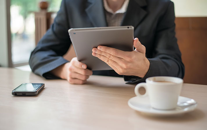 Man sitting at a booth looking at a tablet with a phone and coffee cup on the table.